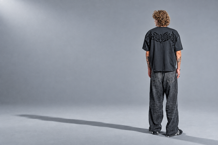 Model seen from behind in a charcoal UNWIND tee and wide grey jeans, standing in a grey studio with a long shadow